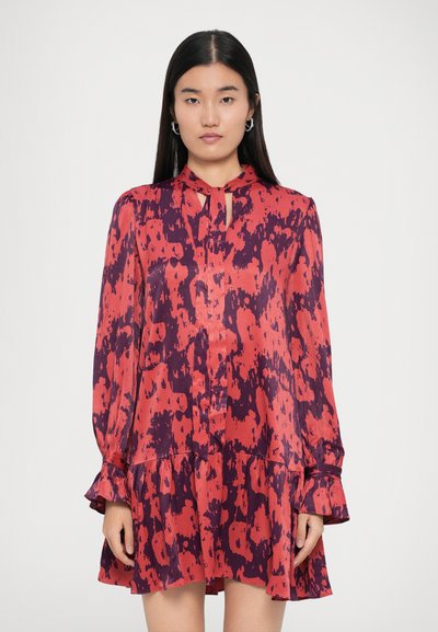 Mujer con cabello largo y oscuro que lleva un vestido de mangas largas de estampado abstracto en rojo y púrpura, con un lazo en el cuello y un dobladillo con volantes, de pie contra un fondo liso.
