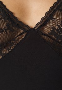 Top en camisole noir avec un décolleté en dentelle ajourée à bords festonnés et un corps lisse. Des panneaux en dentelle texturée mettent en valeur le design.