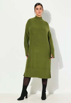 Olivově zelené oversized pletené šaty s vysokým límcem, žebrovanými manžetami, bočními rozparky a dlouhými rukávy, doplněné černými kotníkovými botami.