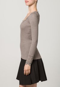 Smeđa rebrasta majica dugih rukava s čipkastim obrubom, uparena s crnom suknjom. Majica ima elegantan izrez i usko kroj.