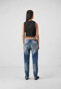 Mujer de espaldas que lleva una parte superior negra sin mangas con cremallera y laterales con cordones, pantalones vaqueros azules descoloridos y zapatos negros, sobre un fondo liso.