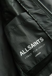 Sort jakke med et netpanel, gittermønster stof og hvid branding med "ALLSAINTS" og "LONDON" trykt på et mærke.