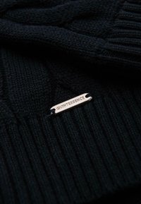 Tissu en maille côtelée noire avec une étiquette rectangulaire en argent gravée du mot "QUINTESSENCE" attachée par des fils noirs.