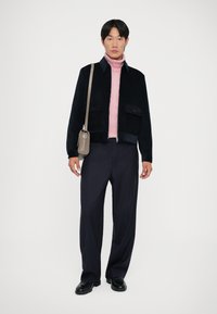 Navy corduroy jacket, pink turtleneck, pinstriped trousers, black shoes, and a small gray bag; modern fit with front pockets and zipper.