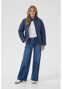 Mornarska puffer jakna s kvadratnim dizajnom, visokim ovratnikom i prednjim zatvaračem, u kombinaciji s širokim plavim trapericama i kremastim cipelama.