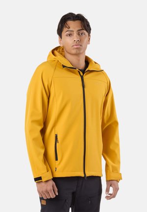 Young man wearing a yellow hooded zip-up jacket and black pants, standing against a plain light background.