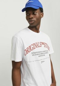 Hvid bomulds t-shirt med rød trykt tekst, rund hals, korte ærmer, med branding og detaljer foran. Blå kasket båret på hovedet.