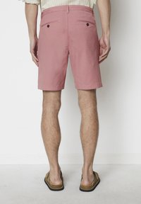 Light pink shorts made of cotton-like material, featuring two back pockets with black button closures, knee-length design, and a smooth texture.
