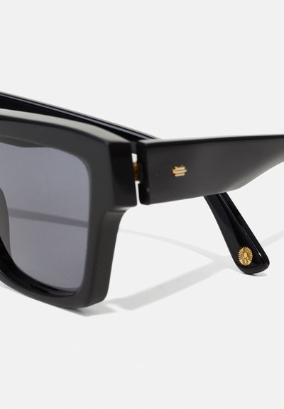 Μαύρα υπερμεγέθη γυαλιά ηλίου με χοντρά πλαίσια, ελαφρώς γυαλιστερή επιφάνεια, γκρίζοι φακοί, χρυσές λεπτομέρειες και ηλιακός μίτος στους κροτάφους.