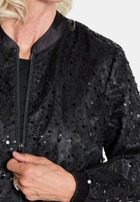 Veste bomber noire avec fermeture éclair, ornée de sequins noirs éparpillés, poignets côtelés et texture satinée.
