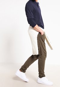 Man som går, iklädd vita sneakers, olivgröna byxor, en marinblå långärmad tröja och en krämfärgad kabelstickad tröja knuten runt midjan.