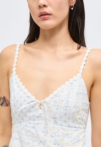 Γυναίκα που φοράει ελαφρύ φλοράλ φόρεμα με λεπτές τιράντες διακοσμημένες με μαργαρίτες και ένα μικρό φιόγκο στο στήθος, που δείχνει ένα πολύχρωμο τατουάζ βουνού στο μπράτσο.