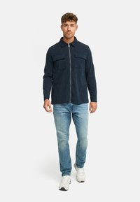 Marineblå zip-up skjorte med to brystlommer, lavet af et tekstureret stof; parret med lysblå jeans og hvide sneakers.