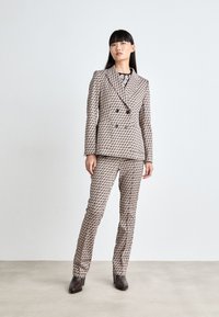 Doppelt geknöpfter Anzug aus gemustertem Stoff in Beige- und Brauntönen. Slim-Fit-Hosen und Blusen sichtbar. Glatte Textur und maßgeschneidertes Design.