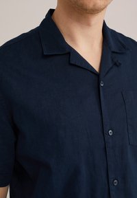 Camisa de manga corta de color azul marino fabricada en lino, con cuello, cierre de botones y un solo bolsillo en el pecho para mayor funcionalidad.
