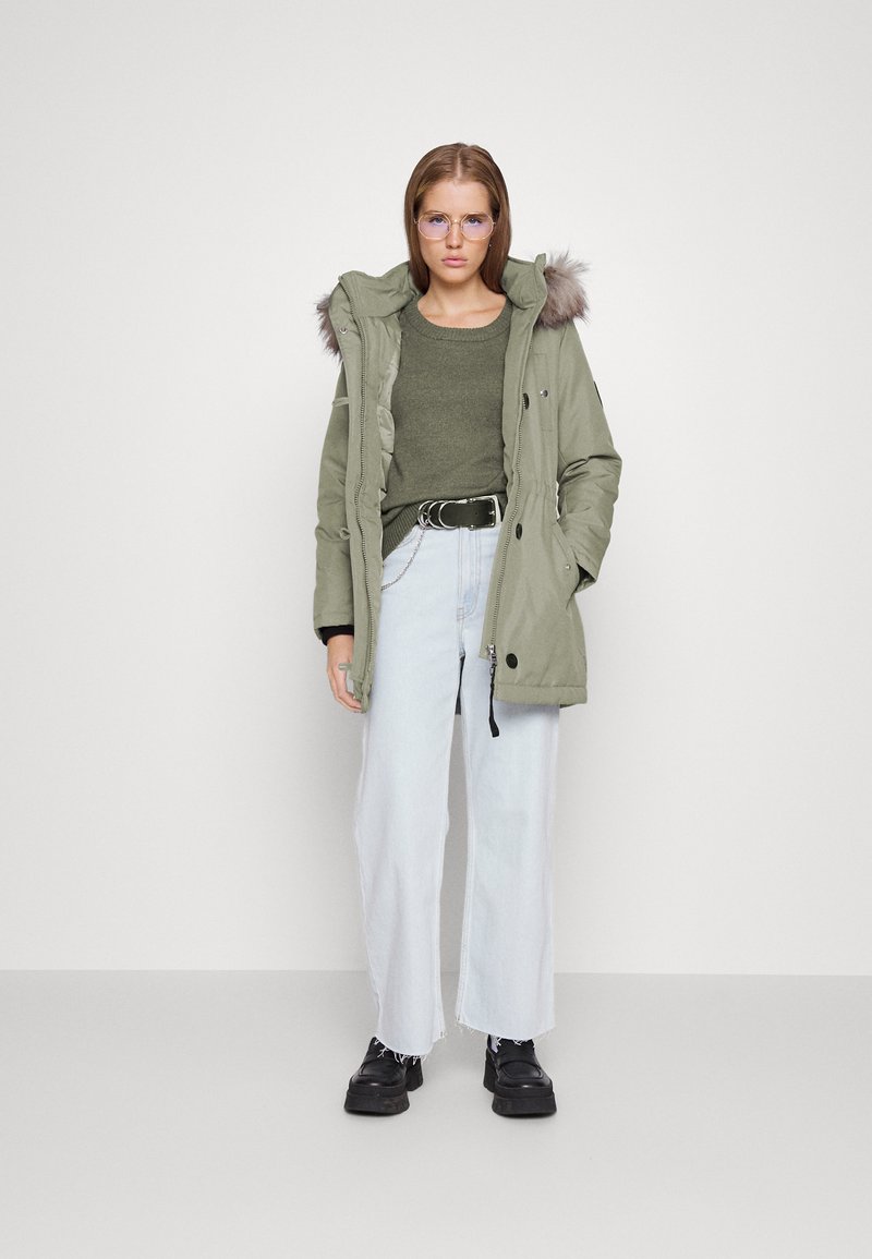 Parka en olive matelassée avec une capuche en fourrure, portée sur un pull vert foncé. Jean large en denim délavé et chaussures noires.