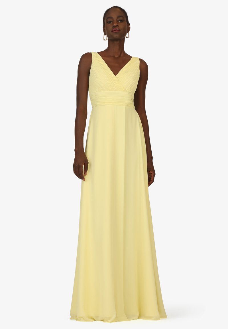 Femme portant une robe jaune sans manches, longueur au sol, avec un décolleté en V et un corsage ajusté et froncé, se tenant devant un fond uni.