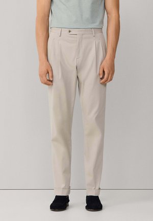 Hombre con pantalones plisados color beige claro con puños remangados y zapatos negros, de pie contra un fondo gris liso.