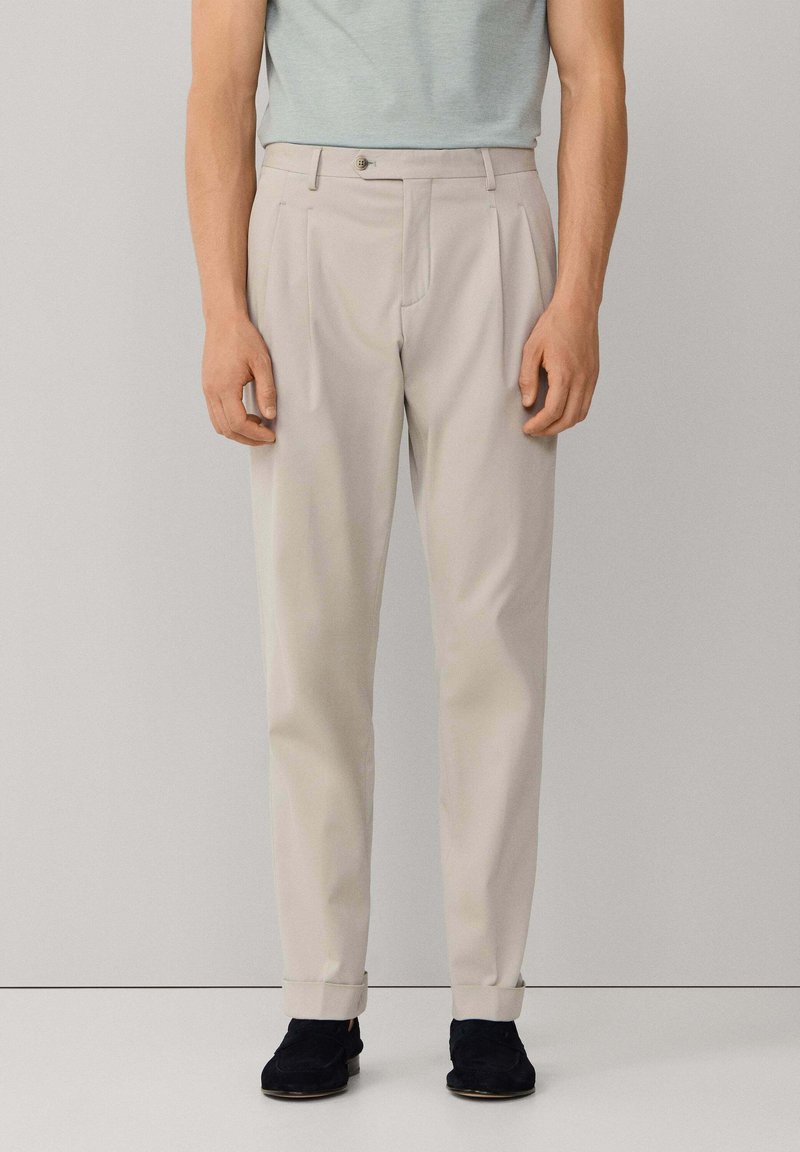 Muškarac u svijetlo bež plisiranim hlačama s pojasnim naborima na rubovima i crnim cipelama, stoji nasuprot običnoj sivoj pozadini.