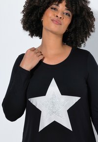 Zwarte langermige top met een grote zilveren sterafbeelding, metallic accenten en een gestructureerd binnenpatroon. Gladde stof met een ronde halslijn.