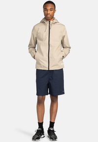 Beige leichte Jacke mit Kapuze, vorderem Reißverschluss und Seitentaschen, kombiniert mit marineblauen Shorts und schwarzen Sneakers. Einfaches und funktionales Design.