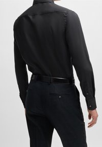 Camicia nera a maniche lunghe con finitura liscia, abbinata a pantaloni scuri con trama. Dettagli notevoli includono una cintura nera lucida e bottoni come accenti.