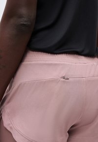 Gros plan d'une personne portant un short de sport rose clair avec une petite poche zippée à l'arrière et un haut noir.