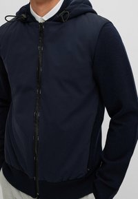 Mörkblå huva jacka med dragkedja fram, strukturerade ärmar och justerbara snören. Tillverkad av en blandning av släta och stickade material.