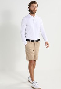 White long-sleeve polo shirt, beige shorts, black belt, and white sneakers with brown accents. Model standing on a light background.