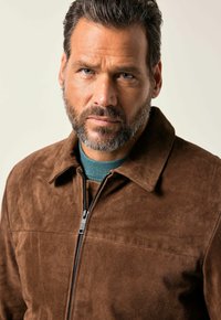 Un hombre con una barba bien cuidada y una expresión intensa lleva una chaqueta de ante marrón sobre un suéter azul, con un fondo claro.