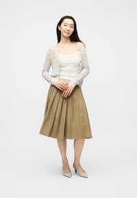 Γυναίκα στέκεται με τα χέρια σταυρωμένα, φοράει ελαφρύ πλεκτό ζακέτα, λευκό πλεκτό τοπ, καφέ πλισέ φούστα και μπεζ τακούνια σε λευκό φόντο.