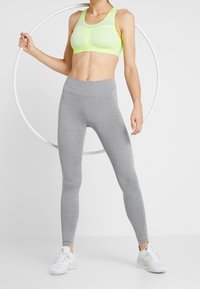Reggiseno sportivo giallo neon con design a costine e spalline larghe, abbinato a leggings grigi aderenti. Il modello tiene un hula hoop bianco.