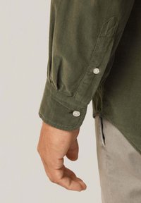 Camisa de pana verde oliva con mangas de puños abotonados. La tela tiene estrías verticales y una textura suave. Presenta un detalle de puño redondeado.