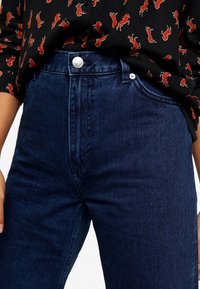 Jeans de mezclilla oscuros de talle alto con una textura suave, que presentan un cierre de botón plateado y bolsillos frontales; combinados con una camisa negra con estampado.