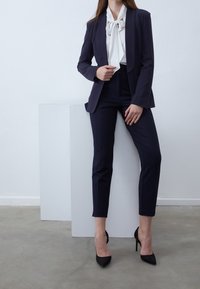 Costume sur mesure bleu marine avec un blazer ajusté et un pantalon slim, associé à un chemisier blanc et des escarpins noirs à bout pointu. Tissu lisse, design minimaliste.
