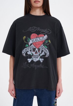 Kvinde iført sort oversized T-shirt med en kranie, korslagte knogler, hjerte og bånd med teksten "Love Kills Slowly" og "Los Angeles".