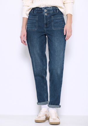 Jeans in denim di lavaggio blu scuro, con vestibilità rilassata, vita elasticizzata, due tasche anteriori e orli arrotolati, abbinati a calzini bianchi e scarpe da ginnastica.