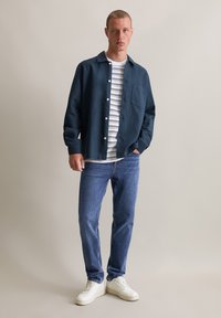 Marineblauw overhemd met knopen over een wit en marineblauw gestreept T-shirt, gecombineerd met blauwe jeans en witte sneakers. Soepele stof en relaxed fit.
