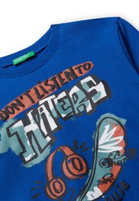 T-shirt en coton bleu avec un col rond et des imprimés graphiques audacieux : "N'écoute pas les haineux" avec des illustrations de skate et de casques audio.