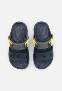 Tummansinisiä sandaaleja, joissa on kuvioitu pohja, säädettävät keltaiset ja harmaat nauhat sekä ilmanvaihtoaukot hengittävyyden lisäämiseksi. Kevyt muotoilu.