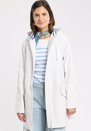 Femme portant un imperméable à capuche blanc sur une chemise rayée bleu et blanc, avec un pantalon à rayures bleu clair et une écharpe bleue autour du cou.