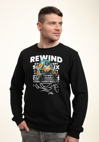 Černá mikina s grafickým potiskem, která zobrazuje ilustrovanou postavu, výrazný text a stylizovaný podpis. Měkký materiál, kulatý výstřih, dlouhé rukávy.