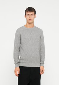 Grå stickad tröja med en strukturerad mönster, rund halsringning och ribbade ärmslut. Kombinerad med svarta byxor står modellen framför en vit bakgrund.