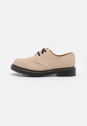 Chaussure beige en daim à lacets avec semelle noire et coutures jaunes, talon bas et bout arrondi, présentée sur fond blanc.