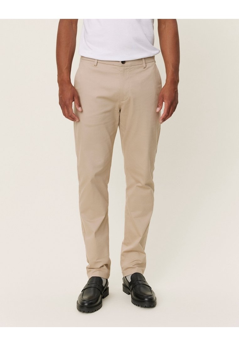 Pantalon beige ajusté en tissu léger, avec poches latérales et taille classique, associé à des mocassins noirs.