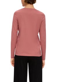 Koszulka z długim rękawem w stonowanym różowym kolorze. Okrągły dekolt i gładka faktura. Prezentowana z tyłu, w połączeniu z czarnymi spodniami.