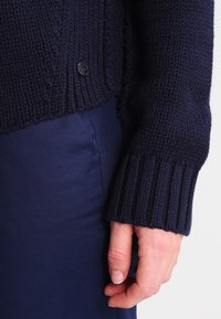 Maglione a maglia blu navy con polsini a coste, caratterizzato da un motivo testurizzato e un piccolo etichetta nera del marchio vicino all'orlo. Abbinato a pantaloni in tessuto blu navy.