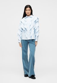 Γυναίκα που φοράει ανοιχτό μπλε πουκάμισο με στρωματοποιημένες βολάν και φαρδιά μπλε τζιν με διακοσμητικές ρίγες στο πλάι, στέκεται με φόντο μια απλή λευκή επιφάνεια.