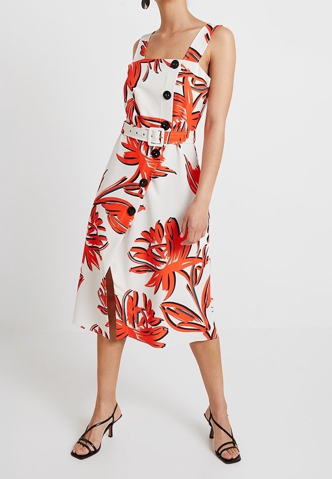 Robe blanche avec un grand imprimé floral rouge, bretelles et boutons noirs. Présente une ceinture et une fente à l'avant. Texture de tissu lisse.