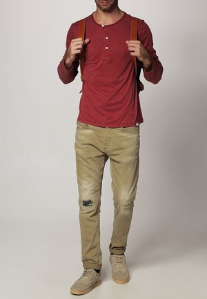 Maglietta henley rossa a maniche lunghe con chiusura a bottoni, abbinata a jeans chiari khaki con effetto strappato e sneakers beige. Spalline in cuoio marrone sulle spalle.